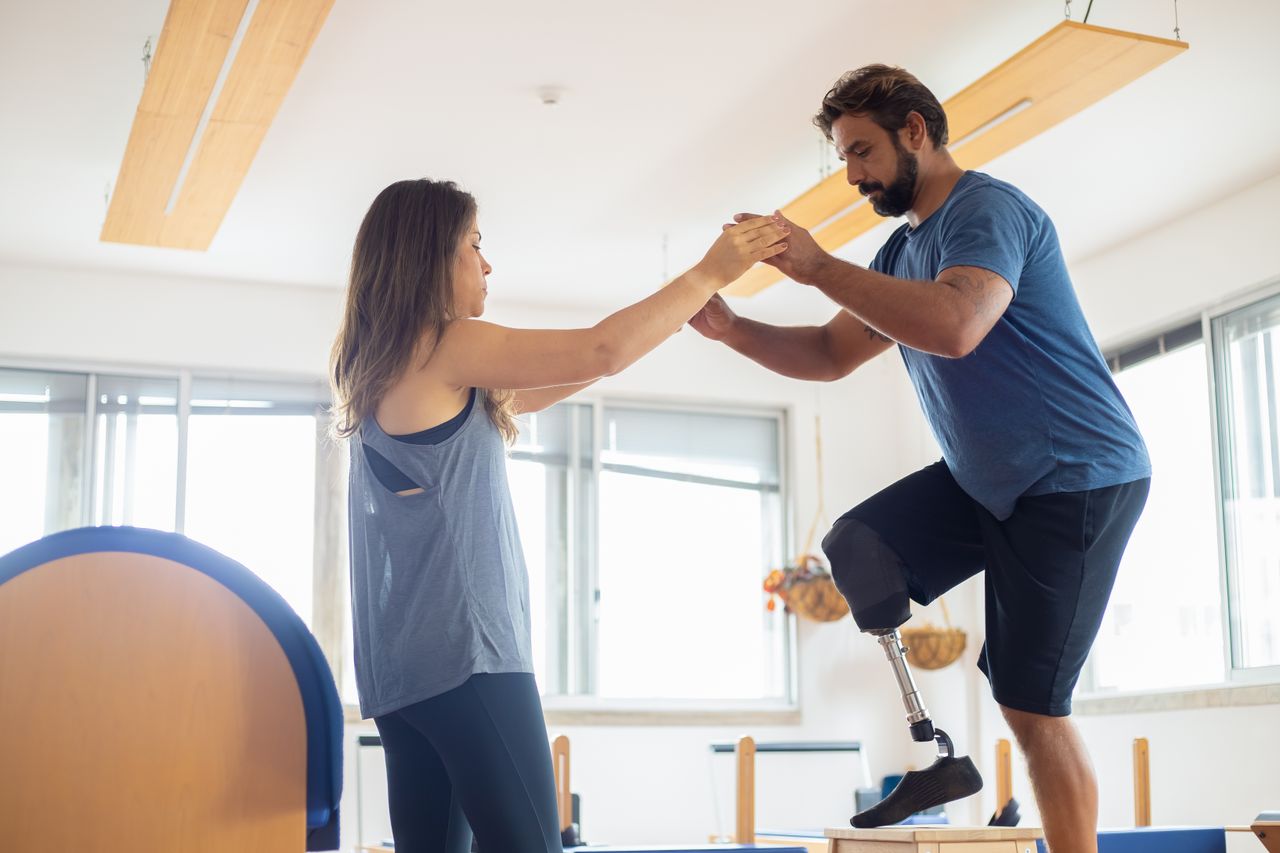 Home A man with a prosthetic leg exercises with a therapist in a well-lit gym environment.